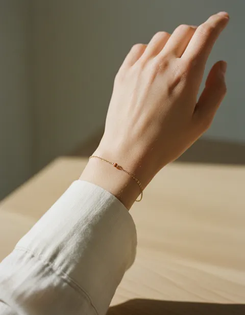 Poignet portant un fin bracelet en chaîne dorée, manche de lin écru, au-dessus d'une table en bois clair